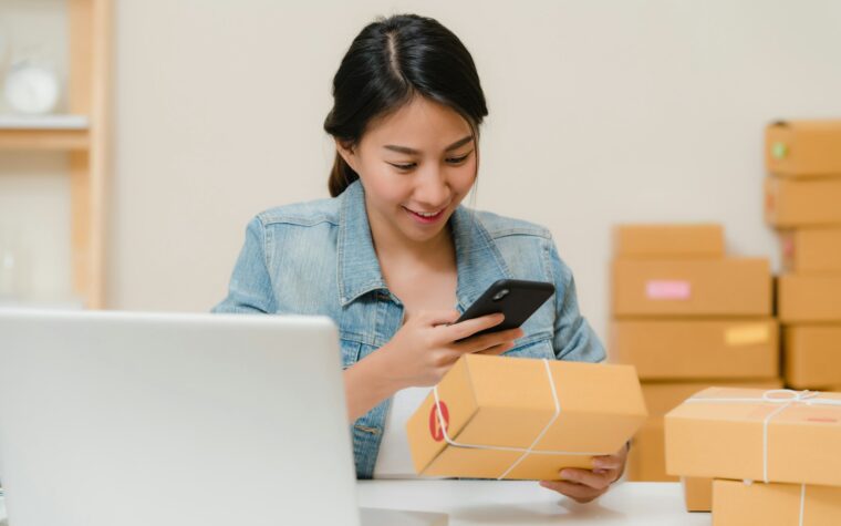 Pemilik usaha kecil memegang HP dan paket, dengan tumpukan kardus pesanan di meja