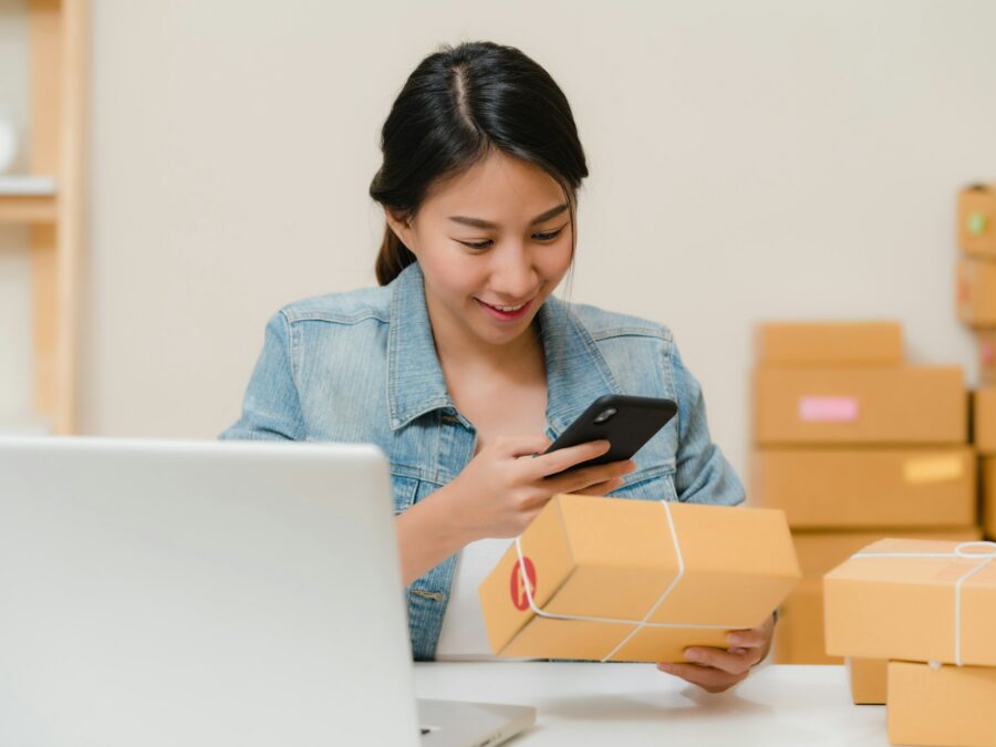 Pemilik usaha kecil memegang HP dan paket, dengan tumpukan kardus pesanan di meja